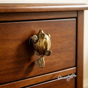 May include: A brass-toned metal cabinet knob shaped like a pufferfish with textured scales, fins, and a tail. The knob is mounted on a dark brown wooden drawer front. The fish figure features a rounded body, prominent eyes, and detailed fins.