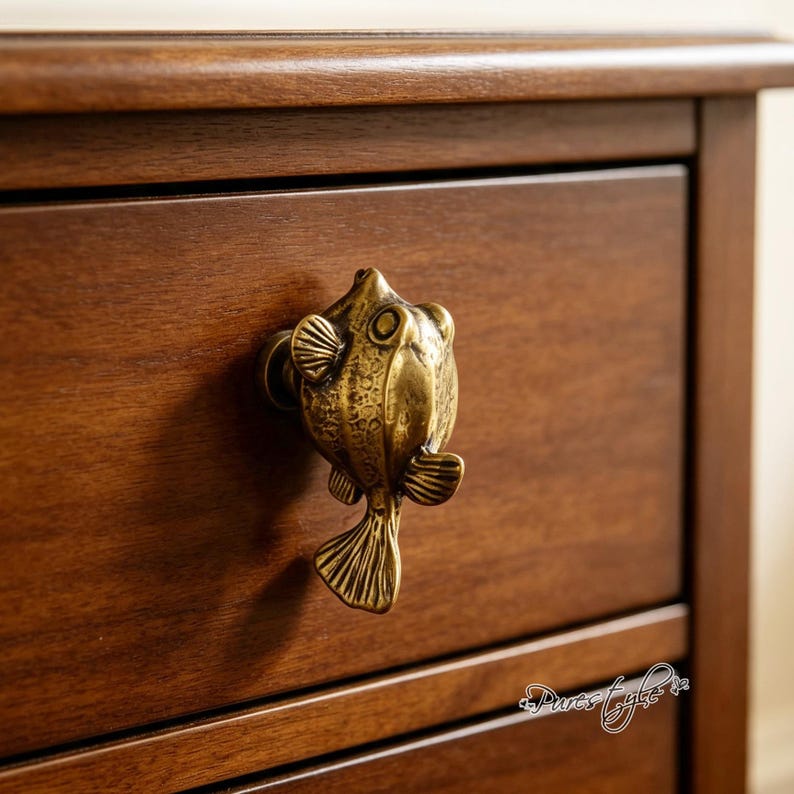 May include: A brass-toned metal cabinet knob shaped like a pufferfish with textured scales, fins, and a tail. The knob is mounted on a dark brown wooden drawer front. The fish figure features a rounded body, prominent eyes, and detailed fins.