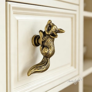 May include: A brass-toned metal drawer knob shaped like a climbing fox with detailed fur texture and a long tail. The fox is mounted to a cream-colored cabinet drawer. The text Purestyle is visible in the bottom right corner of the image.