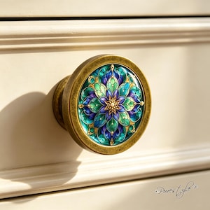 Könnte beinhalten: Ein runder Möbelknopf mit antikem Messing-Finish und einer zentralen kreisförmigen Einlage, die ein lebendiges Mandala-Muster in Türkis-, Smaragdgrün- und tiefen Violetttönen mit goldenen Akzenten auf einer glatten, cremefarbenen Oberfläche zeigt.