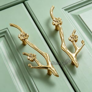 May include: Two gold-colored cabinet handles in a branch and flower design. The handles are mounted on a light green cabinet door. The handles have a curved branch shape with small flower details. The handles are made of metal.