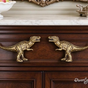 May include: Two metallic gold-toned dinosaur-shaped drawer pulls mounted on a dark wood cabinet. Each pull is shaped like a Tyrannosaurus Rex with detailed scales and texture, facing inward toward each other. The hardware is attached to the front.