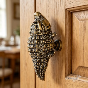 May include: A brass-toned metal door knob shaped like an alligator with a textured, scaly surface and a dark finish in the crevices. The knob is mounted on a light-colored wooden door panel and features a small, round base connecting it to the wood.
