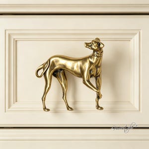 Peut inclure: Une poignée de meuble en métal doré en forme de lévrier debout. Le chien est représenté de profil, la tête légèrement tournée vers le haut et une patte avant levée. La surface présente une finition polie avec des détails sombres en creux.