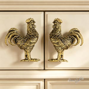 May include: Two antique brass-toned metal cabinet knobs shaped like roosters. Each knob features detailed plumage, a comb, and a wattle, with a metallic finish that highlights the textured feathers. The roosters are facing each other.