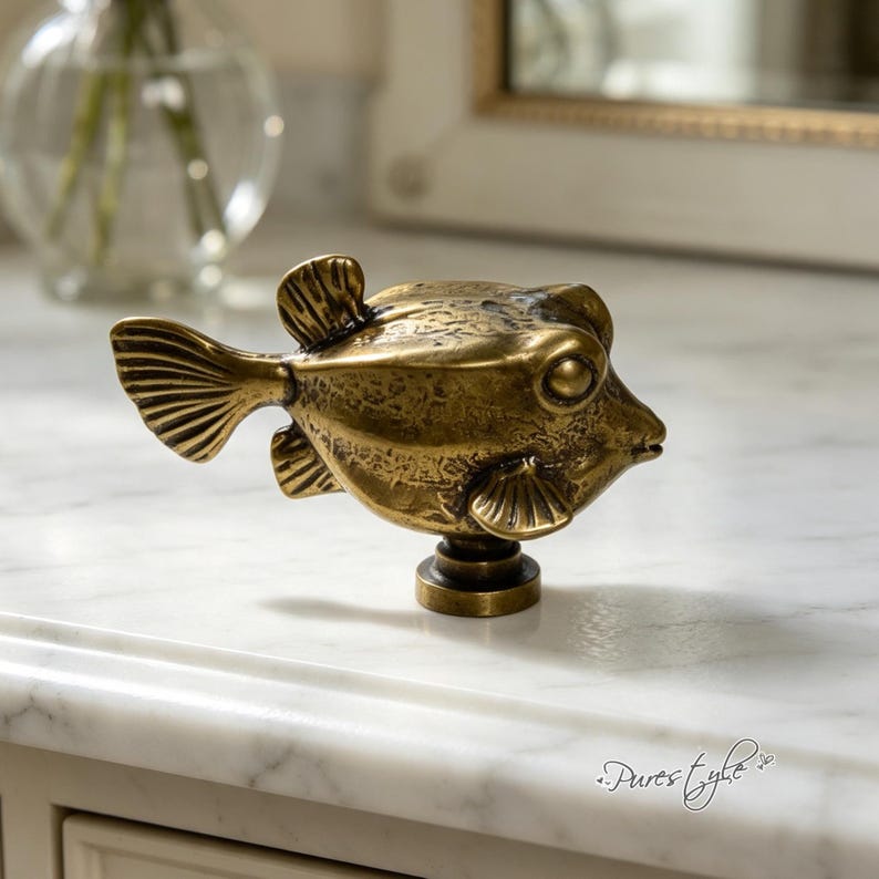 May include: A brass-toned metal cabinet knob shaped like a pufferfish. The knob features a textured, weathered surface with detailed fins, a prominent eye, and a small mouth. It is mounted on a circular base, showcasing a metallic finish.