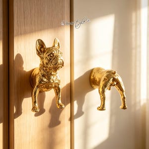 May include: Two gold-colored metal cabinet knobs shaped like a French bulldog. One knob depicts the front of the dog, and the other depicts the rear of the dog. The text "Purestyle" is visible above the front-facing knob on the wooden surface.