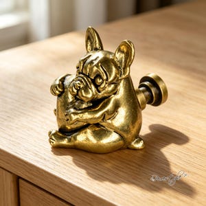 May include: A brass-toned metal cabinet knob shaped like a French bulldog sitting and holding a round object. The knob features a detailed, textured finish with dark highlights in the crevices and a cylindrical mounting post extending from the back.