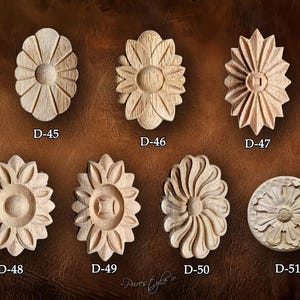 Puede incluir: Una colección de siete tallas florales de madera sin terminar, cada una con un diseño único. Las tallas son ovaladas y circulares, con varios detalles de pétalos y centro. Cada talla está etiquetada con un número "D-".