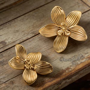 Puede incluir: Dos tiradores de armario dorados con forma de flor. Cada tirador tiene cinco pétalos con vetas detalladas y un pequeño diseño floral central. Los tiradores están colocados sobre una superficie de madera marrón claro envejecida.
