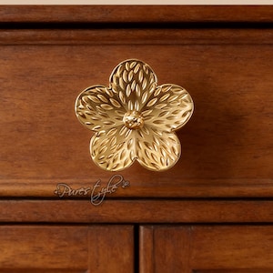 Könnte beinhalten: Ein goldfarbener Schrankknauf in Blumenform. Der Knauf ist an einem dunkelbraunen Holzschrank befestigt. Das Blumendesign hat detaillierte Blütenblattstrukturen und einen zentralen, abgerundeten Akzent. Der Schrank hat zwei Türen.