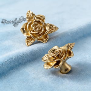 May include: Two gold-colored rose-shaped cabinet knobs. Each knob features detailed petals and leaves, with a stem for mounting. The knobs are displayed on a light blue, velvety surface. The knobs are likely made of metal.