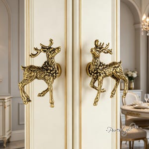 May include: Two antique brass-colored metal door handles shaped like standing deer with detailed antlers and textured fur. The handles are mounted on a white double door with gold trim, featuring a sculptural design that mimics the form of a stag.