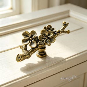 Puede incluir: Un tirador de mueble de metal en tono latón con forma de rama de árbol con dos flores prominentes. El herraje presenta un acabado antiguo texturizado con pétalos detallados y una base de montaje central. El texto Purestyle es visible en la esquina.