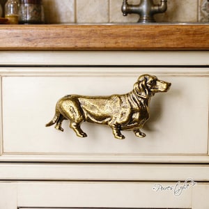 May include: A brass-toned metal cabinet pull shaped like a standing dachshund dog. The hardware features a textured, antique-style finish with detailed sculpting of the dog's fur, ears, and facial features, mounted horizontally on a drawer front.