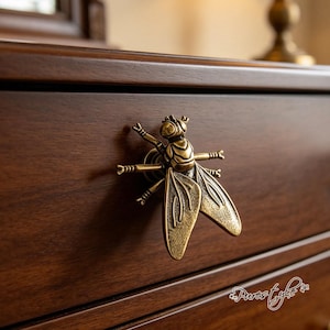 May include: A brass-toned metal cabinet knob shaped like a bee with detailed wings and legs. The insect-shaped hardware is mounted onto a dark brown wooden drawer front. The design features a textured finish on the wings and a rounded head.