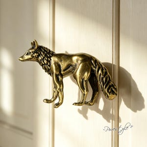 May include: A brass-toned metal cabinet knob shaped like a standing fox. The fox figure features detailed fur textures, pointed ears, and a bushy tail, with a metallic finish that reflects light, creating a distinct shadow on the surface behind it.