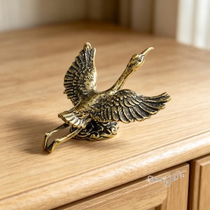 May include: A brass-coloured metal figurine of a bird in flight with wings spread wide. The bird features detailed feather textures and is mounted on a small, rounded base with a swirling pattern. The finish has a dark, antiqued patina.