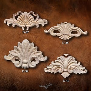 Puede incluir: Cuatro tallas decorativas de madera sin terminar en varios diseños florales y de volutas. Cada talla está etiquetada con un número "D-". Las tallas se muestran sobre un fondo marrón. Las tallas son probablemente para muebles o decoración del hogar.