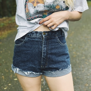 May include: A woman wearing a light gray t-shirt with a multi-colored print of foxes and wolves. She is also wearing dark blue denim shorts with a frayed hem.