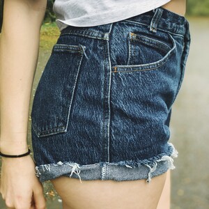 May include: A pair of blue denim shorts with a frayed hem and a cuffed leg. The shorts have a back pocket and are worn by a person.