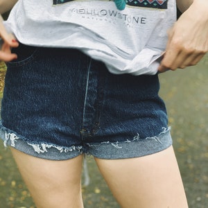 May include: A person wearing a light gray t-shirt with a graphic print of a forest scene and the text "Yellowstone National Park". The person is also wearing dark blue denim shorts with a frayed hem.