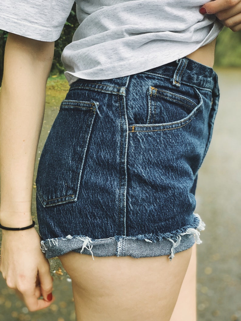 May include: A pair of blue denim shorts with a frayed hem and a cuffed leg. The shorts have a back pocket and are worn by a person.