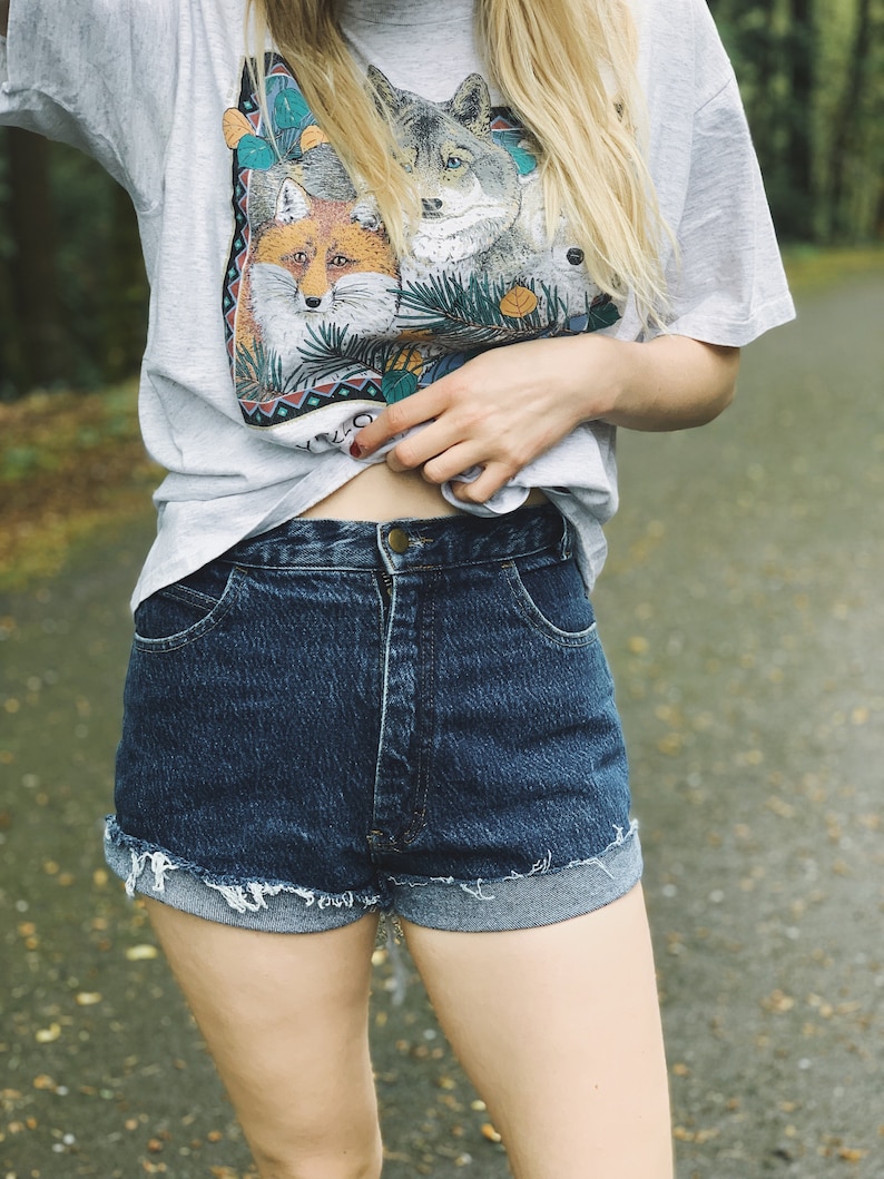 May include: A woman wearing a light gray t-shirt with a multi-colored print of foxes and wolves. She is also wearing dark blue denim shorts with a frayed hem.
