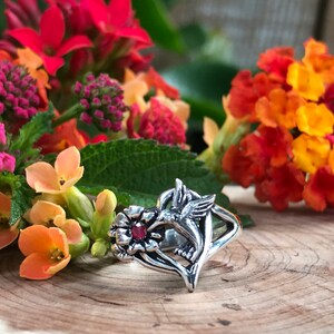 May include: A silver ring featuring a hummingbird with a red gemstone in its beak, surrounded by a stylized flower design.