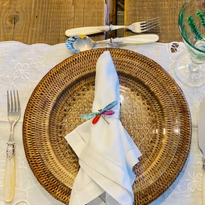 Puede incluir: Una servilleta blanca con un anillo de servilleta de libélula en un plato de servicio de mimbre marrón. El plato de servicio está sobre un mantel de encaje blanco. Hay un tenedor y un cuchillo en la mesa.