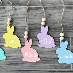 May include: Five pastel colored bunny shaped wooden ornaments with names written on them. The bunnies are hanging from twine with wooden beads. The names are Lucas, James, Sophia, Olivia, and Noah.