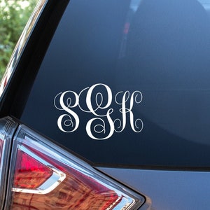 May include: A white monogram decal with the letters "SOK" in a cursive font on the back window of a gray car.