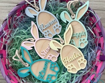 May include: Five wooden bunny-shaped Easter egg ornaments with personalized names. The ornaments are painted in pastel colors, including pink, blue, yellow, and green. The names on the ornaments are Lauren, Stevie, Maggie, Hailee, and Liam.
