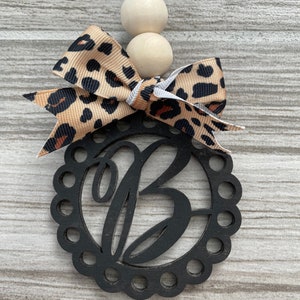 May include: Black wooden ornament with a scalloped edge and the letter "B" cut out. The ornament is decorated with a brown and black leopard print bow and white wooden beads.
