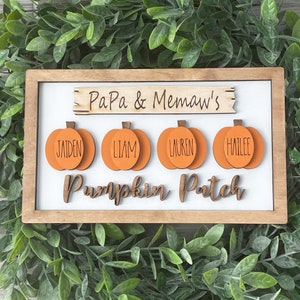 Puede incluir: Un letrero enmarcado en madera con fondo blanco. El letrero presenta recortes de calabazas naranjas con nombres, y las palabras "Papa & Memaw's" y "Pumpkin Patch" en letras de madera. El letrero está rodeado de vegetación.