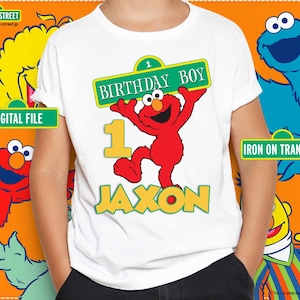 May include: White t-shirt featuring a red Elmo character holding a sign that reads "BIRTHDAY BOY" above the name "JAXON" in yellow. The number "1" is also displayed. The shirt is a digital file or iron-on transfer.