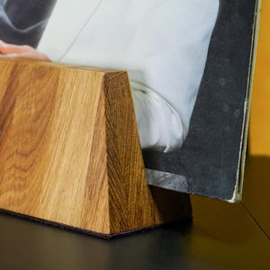 Könnte beinhalten: Ein hölzerner Schallplattenhalter in Trapezform, der eine Schallplatte präsentiert. Der Halter ist aus hellbraunem Holz gefertigt und steht auf einer dunklen Oberfläche. Das Plattencover zeigt eine Person in einem weißen Jackett.
