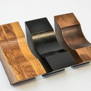 May include: Three wooden decorative shelf brackets in varying finishes: natural wood, black, and dark brown. Each bracket has a curved design and a rectangular base. The brackets are displayed on a white background.