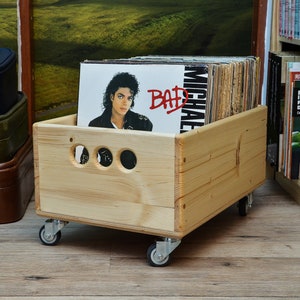 Puede incluir: Una caja de discos de madera sobre ruedas, llena de discos de vinilo. La caja tiene tres recortes circulares en el lateral. La portada del álbum visible presenta a Michael Jackson y la palabra "BAD". Maletas vintage apiladas en el fondo.