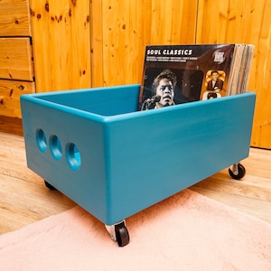 Może przedstawiać: Drewniana skrzynia do przechowywania płyt winylowych w kolorze turkusowym z kółkami. Skrzynia ma trzy wycięte koła po bokach i jest wypełniona płytami winylowymi. Płyta na górze ma napis "SOUL CLASSICS" na okładce.