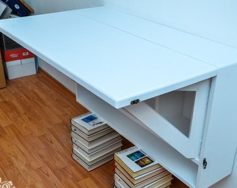 May include: A white, wall-mounted desk with a hinged tabletop. The desk is supported by a white frame and a shelf. Books are stacked beneath the desk. The desk is mounted on a white wall and the floor is wood.