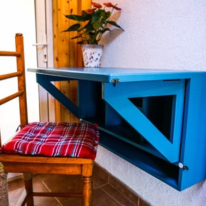 May include: A blue folding wall-mounted desk with a wooden chair and a red and white plaid cushion. The desk has a shelf underneath and is mounted to a white wall.