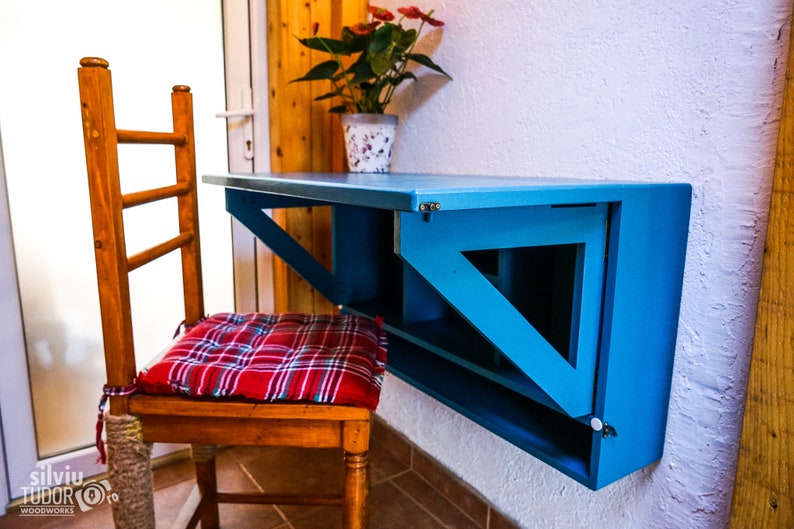 May include: A blue folding wall-mounted desk with a wooden chair and a red and white plaid cushion. The desk has a shelf underneath and is mounted to a white wall.
