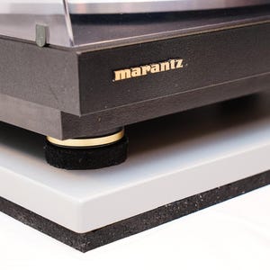 May include: Close-up of a black Marantz turntable with gold lettering. The turntable sits on a gray platform with black rubber feet. The image is well-lit, highlighting the details of the audio equipment.