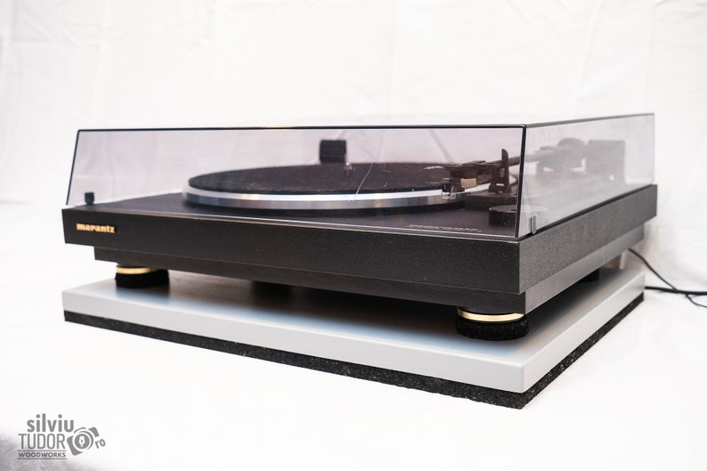 May include: A black Marantz turntable with a clear dust cover and a silver platter. The turntable is sitting on a gray platform with gold feet.