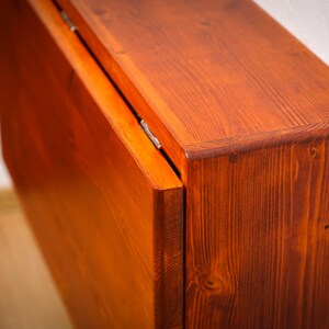 May include: A wooden folding table with a rich, reddish-brown stain. The table is closed and the top is folded down, revealing the hinges and the grain of the wood.
