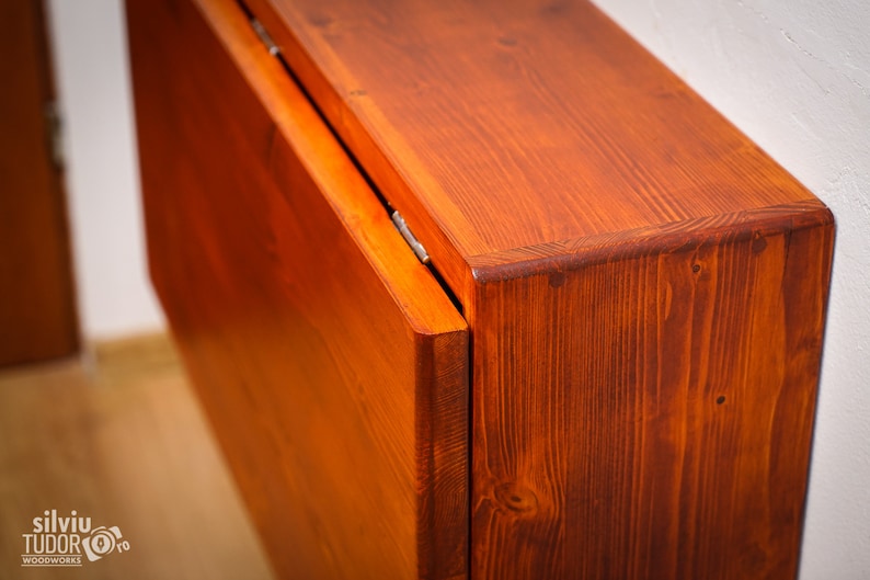 May include: A wooden folding table with a rich, reddish-brown stain. The table is closed and the top is folded down, revealing the hinges and the grain of the wood.