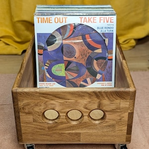 Puede incluir: Caja de almacenamiento de discos de vinilo de madera sobre ruedas, llena de vinilos. La caja es de madera marrón claro con tres recortes circulares en la parte delantera. Las fundas de los discos muestran los títulos "TIME OUT" y "TAKE FIVE".