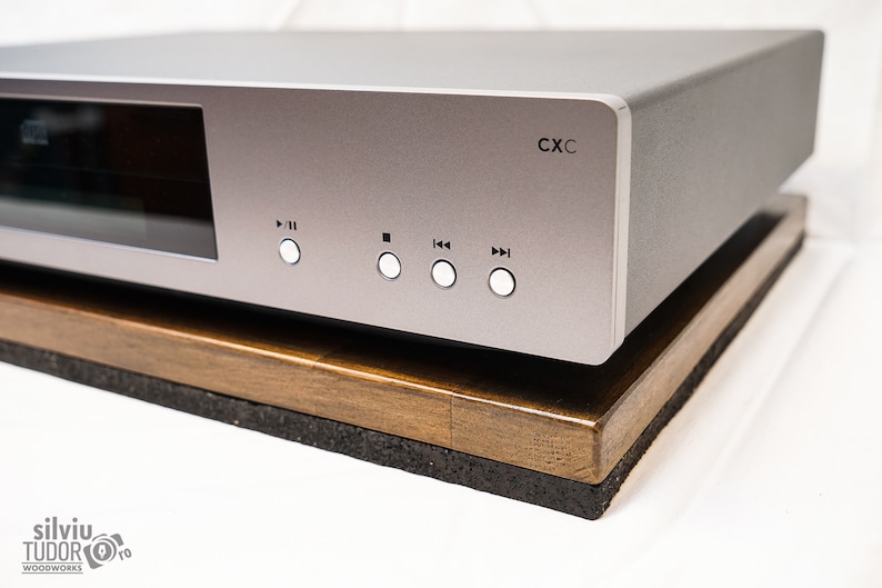 May include: A silver audio component with a black display and white buttons labeled "Play/Pause", "Stop", "Previous", and "Next". The component is sitting on a wooden stand with a black base.