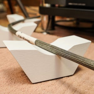 May include: A close-up of a gray and black braided cable resting on two white, geometric cable supports. The supports are on a light brown surface. The cable has a white label with text.